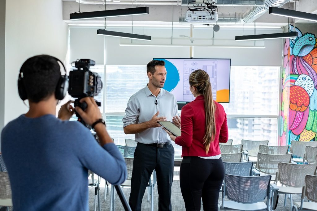 Happy businessman during corporate interview with female journalist. Manager answering question in office. Young woman at work as reporter with business man and cameraman shooting video for broadcast